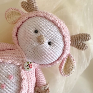 Puede incluir: Un juguete de reno de crochet rosa y blanco con un bot&oacute;n floral y cuernos marrones. El juguete lleva una chaqueta de crochet rosa.