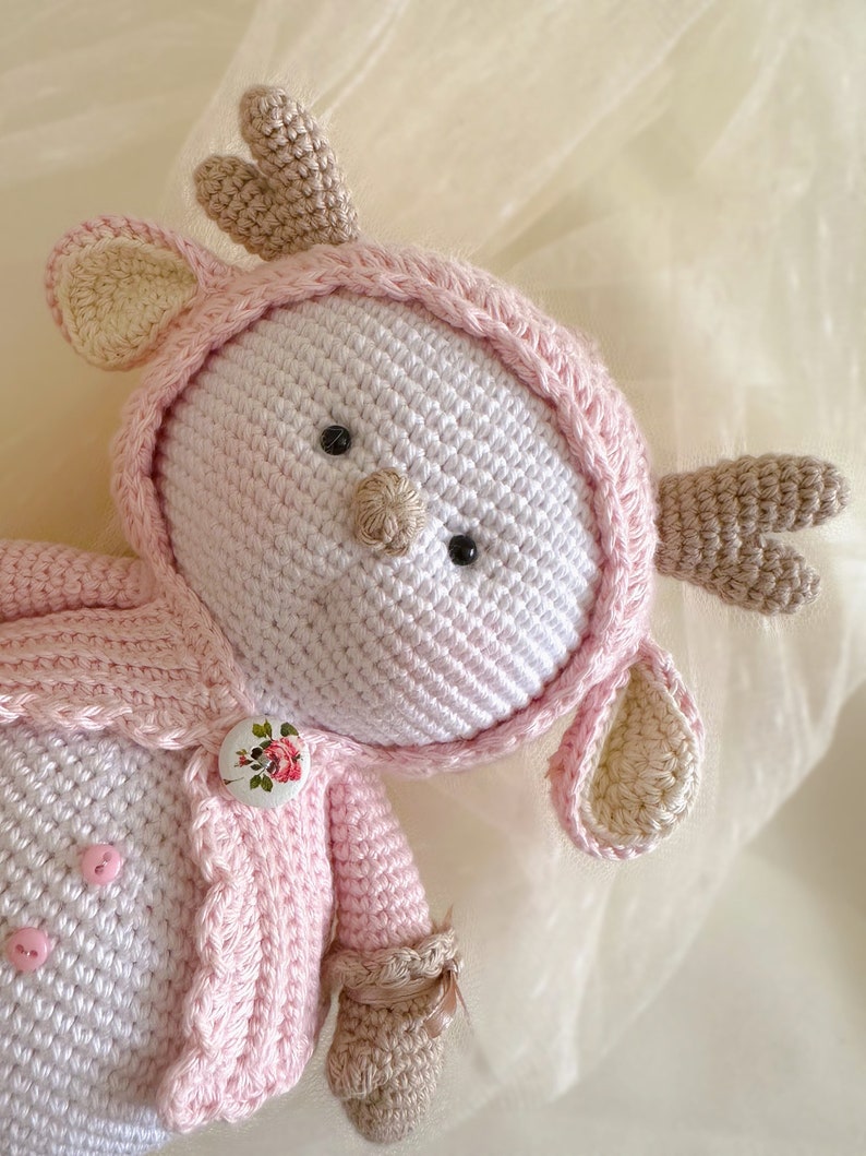 Puede incluir: Un juguete de reno de crochet rosa y blanco con un bot&oacute;n floral y cuernos marrones. El juguete lleva una chaqueta de crochet rosa.