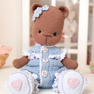 Op de afbeelding: Een handgemaakte gehaakte teddybeerpop. De beer is bruin en draagt een blauwe jurk met witte accenten. De jurk heeft bloemenknopen en witte kanten afwerking. De voeten van de beer hebben een roze hartontwerp. Een blauwe bloem siert het hoofd van de beer.