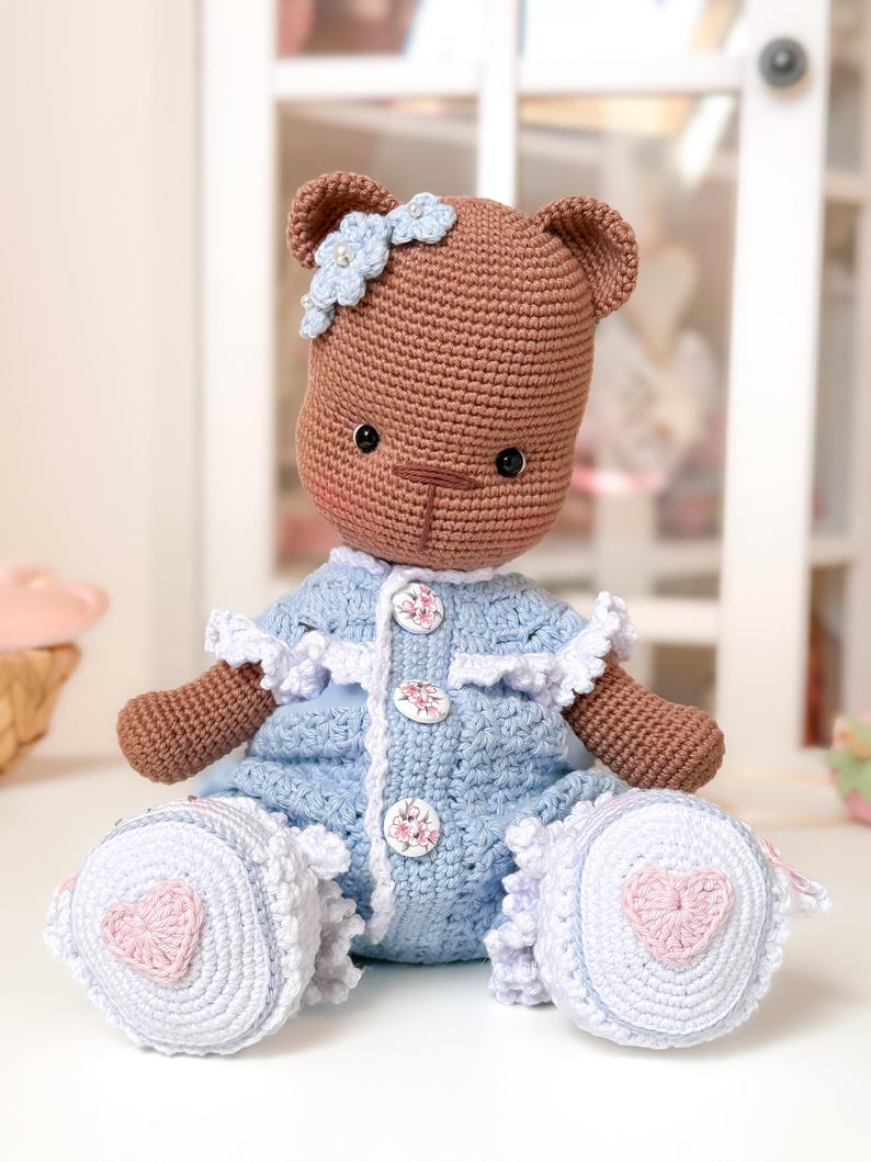 Op de afbeelding: Een handgemaakte gehaakte teddybeerpop. De beer is bruin en draagt een blauwe jurk met witte accenten. De jurk heeft bloemenknopen en witte kanten afwerking. De voeten van de beer hebben een roze hartontwerp. Een blauwe bloem siert het hoofd van de beer.