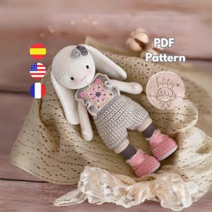 Puede incluir: Una muñeca de conejo de ganchillo hecha a mano con cuerpo blanco, orejas largas y un atuendo gris y rosa. La muñeca tiene un diseño floral en el pecho y lleva botas rosas. La imagen incluye el texto "PDF Pattern" y un logotipo.