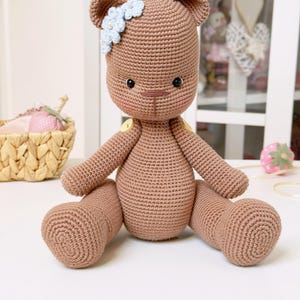 Op de afbeelding: Een handgemaakte bruine gehaakte teddybeer met een lichtblauwe bloem haaraccessoire. De beer heeft zwarte veiligheidsoogjes, een kleine bruine neus en gele knoopaccenten. De beer zit met zijn benen uitgestrekt.