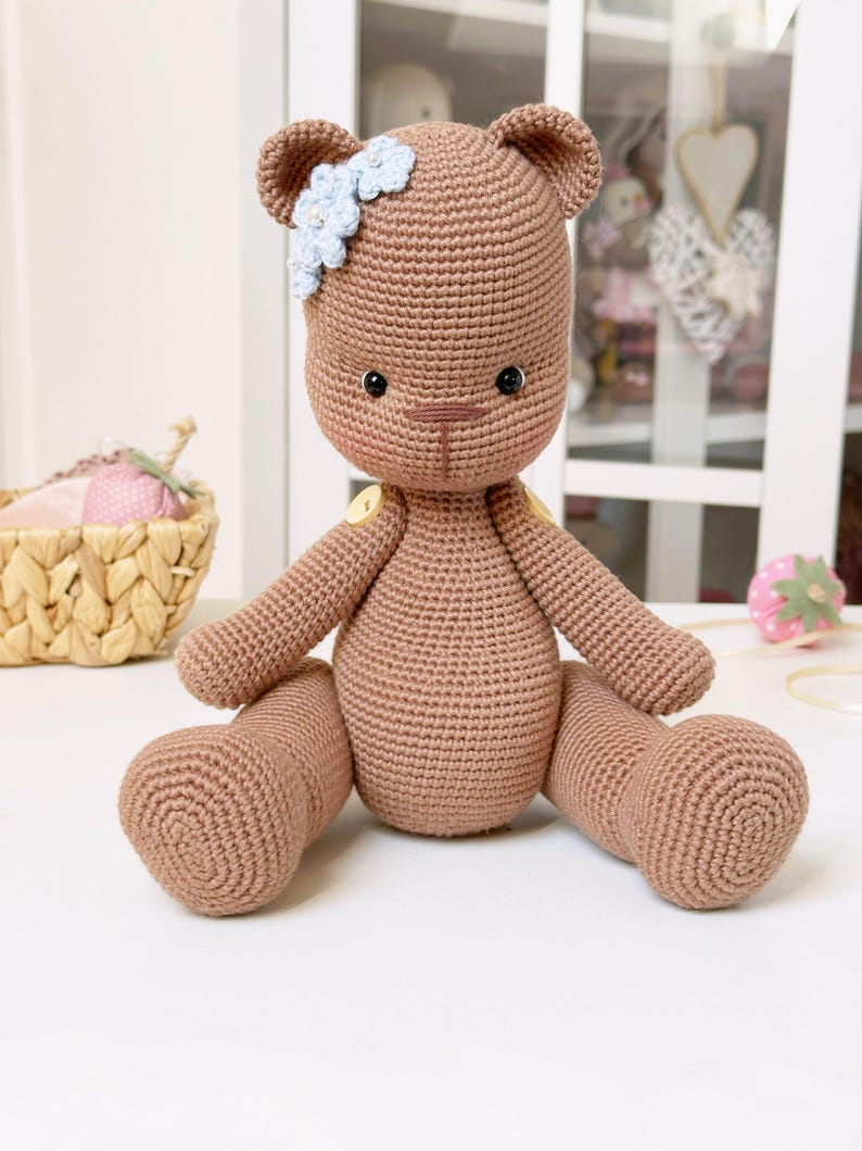 Op de afbeelding: Een handgemaakte bruine gehaakte teddybeer met een lichtblauwe bloem haaraccessoire. De beer heeft zwarte veiligheidsoogjes, een kleine bruine neus en gele knoopaccenten. De beer zit met zijn benen uitgestrekt.