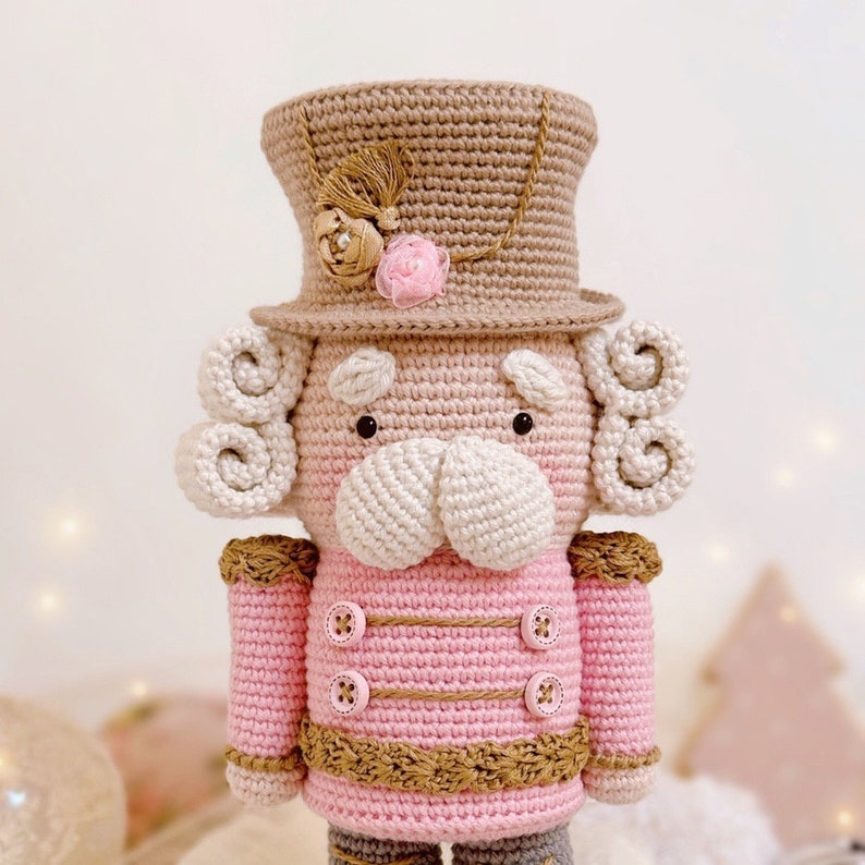 May include: A crocheted nutcracker doll with a pink and brown outfit, a white beard, and a brown top hat with a pink flower.