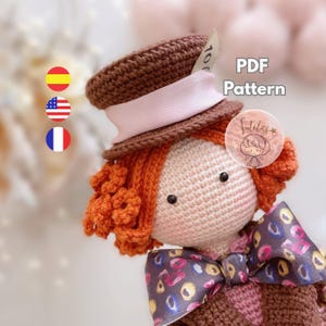 Peut inclure: Une poupée crochetée avec des cheveux bouclés orange, un chapeau haut de forme marron avec un ruban rose et un nœud papillon à motifs. La poupée a un visage rose et porte une tenue marron et rose. Le texte "PDF Pattern" est visible.