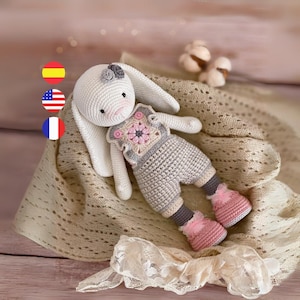 Puede incluir: Un juguete de conejo de crochet que lleva un atuendo de crochet gris y rosa con un diseño de flores. El conejo está acostado sobre una manta de crochet beige.
