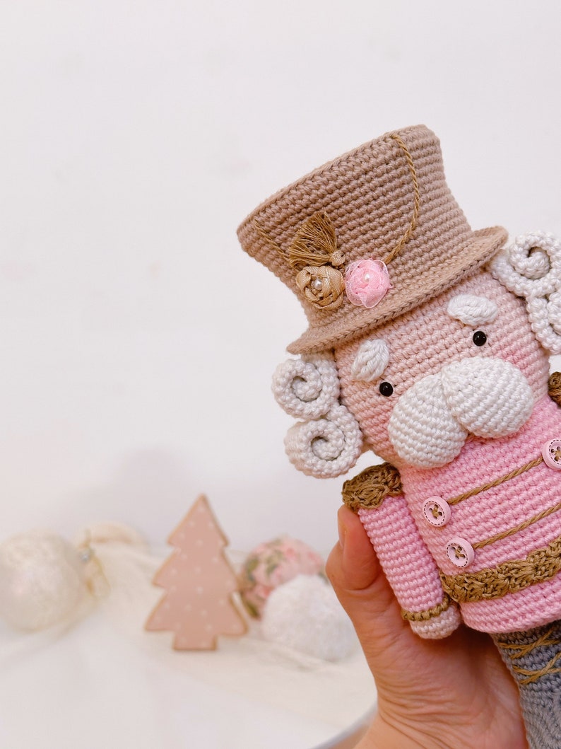 May include: A crocheted nutcracker doll with a pink and white outfit, a brown hat with a pink flower, and a white beard. The doll is being held by a hand.