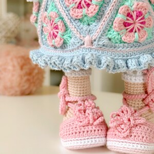 K&ouml;nnte beinhalten: Eine geh&auml;kelte Puppe mit einem blauen und gr&uuml;nen Kleid mit rosa Blumen. Die Puppe tr&auml;gt rosa Schuhe mit wei&szlig;en Verzierungen.