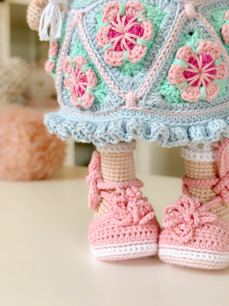 K&ouml;nnte beinhalten: Eine geh&auml;kelte Puppe mit einem blauen und gr&uuml;nen Kleid mit rosa Blumen. Die Puppe tr&auml;gt rosa Schuhe mit wei&szlig;en Verzierungen.