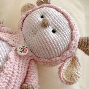 Puede incluir: Un juguete de reno de crochet con una chaqueta de punto rosa y un bot&oacute;n blanco con un dise&ntilde;o floral. El reno tiene una cara blanca con ojos negros y una nariz marr&oacute;n.