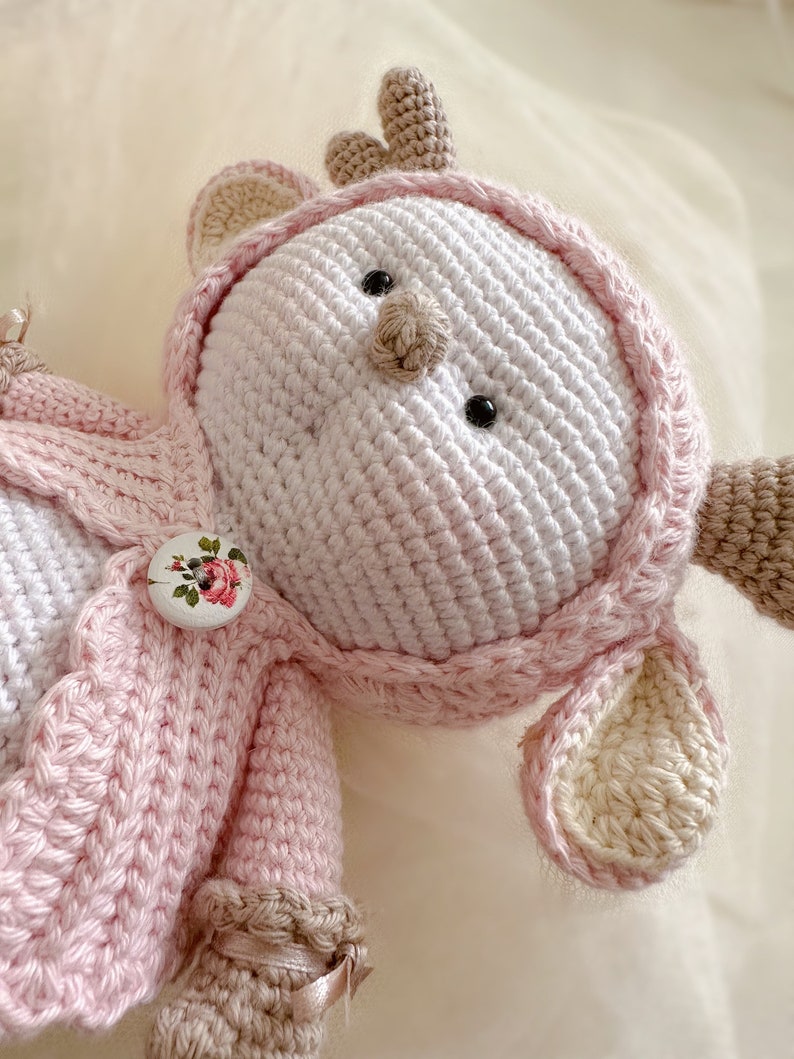 Puede incluir: Un juguete de reno de crochet con una chaqueta de punto rosa y un bot&oacute;n blanco con un dise&ntilde;o floral. El reno tiene una cara blanca con ojos negros y una nariz marr&oacute;n.