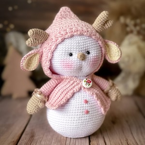 Puede incluir: Un mu&ntilde;eco de nieve de crochet que lleva un su&eacute;ter rosa con capucha y cuernos. El mu&ntilde;eco de nieve tiene un bot&oacute;n y dos puntos blancos en su cuerpo.