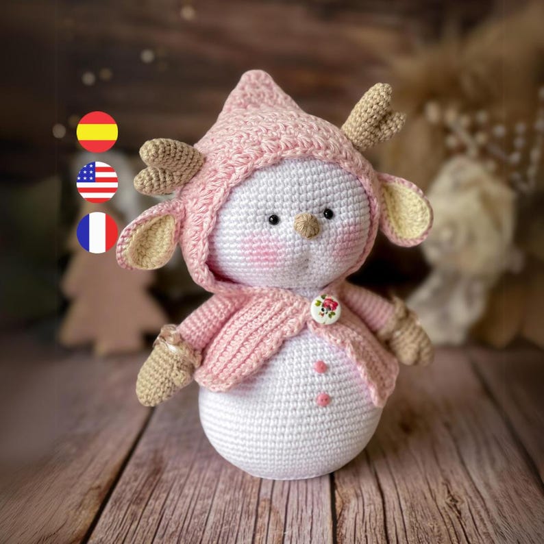 Puede incluir: Un mu&ntilde;eco de nieve de crochet que lleva un su&eacute;ter rosa con capucha y cuernos. El mu&ntilde;eco de nieve tiene un bot&oacute;n y dos puntos rosas en su cuerpo.