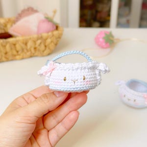 Op de afbeelding: Een kleine, witte gehaakte tas met een lichtblauw handvat en een roze strik. De tas heeft een schapenachtig gezicht met roze wangen en kleine oren. Een tweede, vergelijkbare tas is zichtbaar. De tas wordt in een hand gehouden.
