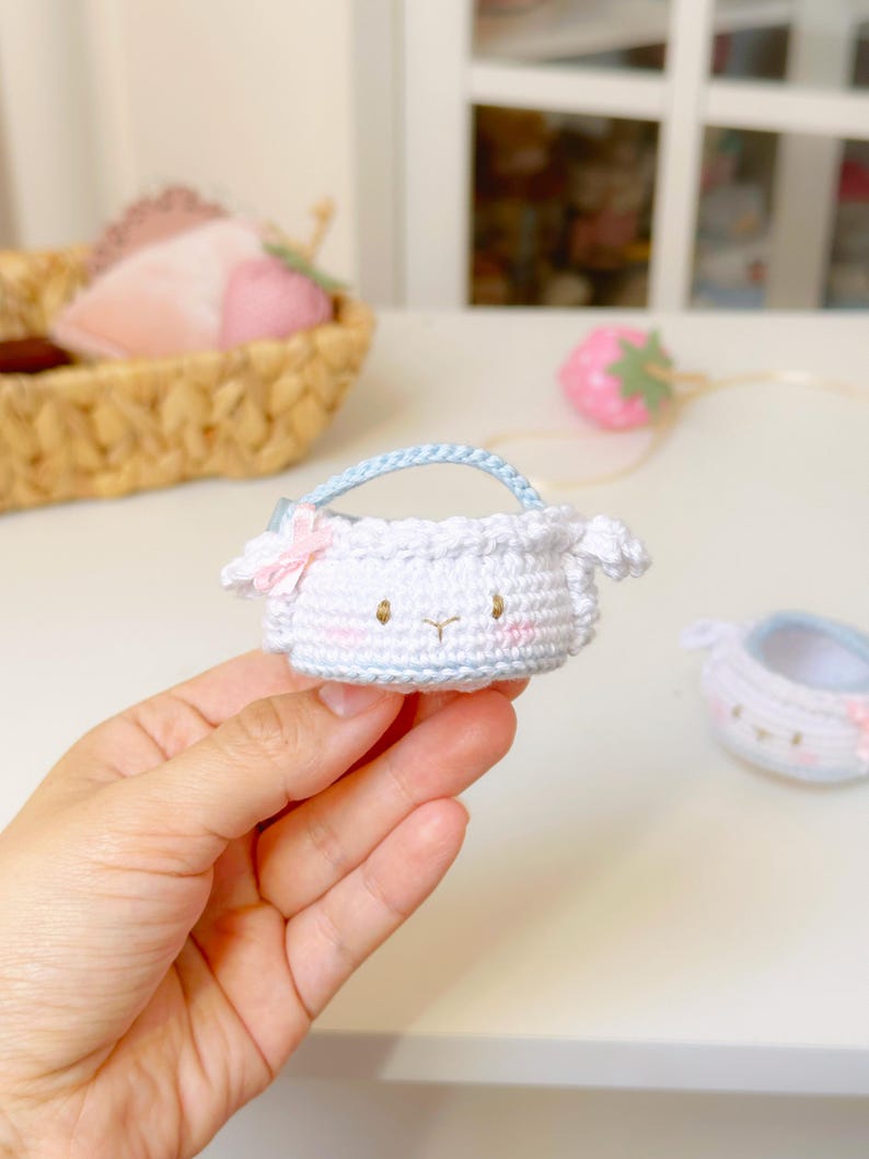 Op de afbeelding: Een kleine, witte gehaakte tas met een lichtblauw handvat en een roze strik. De tas heeft een schapenachtig gezicht met roze wangen en kleine oren. Een tweede, vergelijkbare tas is zichtbaar. De tas wordt in een hand gehouden.