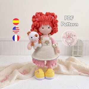 Puede incluir: Una muñeca de ganchillo con pelo rizado naranja vibrante, vestida con un pichi color crema con un pequeño bordado floral, pantalones rosas con volantes y zapatos amarillos. La muñeca sostiene un pequeño gato de ganchillo blanco y marrón. Texto PDF Pattern.
