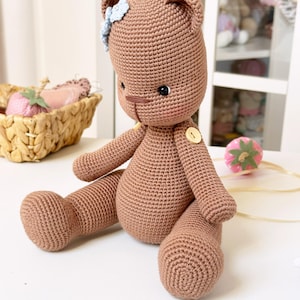 Op de afbeelding: Een handgemaakte, gehaakte teddybeer in een bruine kleur. De beer heeft een blauwe bloem op zijn hoofd en een knoopdetail op zijn borst. De beer zit met zijn benen uitgestrekt, wat de gedetailleerde haaktextuur laat zien.