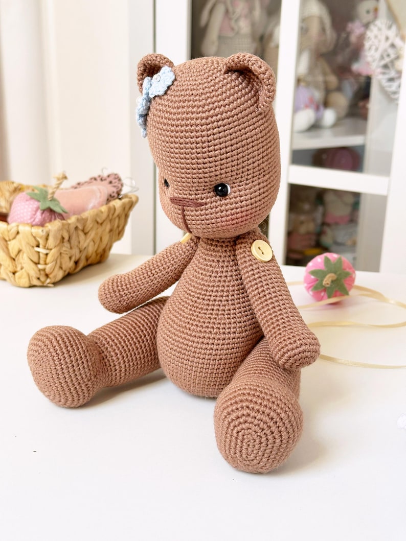 Op de afbeelding: Een handgemaakte, gehaakte teddybeer in een bruine kleur. De beer heeft een blauwe bloem op zijn hoofd en een knoopdetail op zijn borst. De beer zit met zijn benen uitgestrekt, wat de gedetailleerde haaktextuur laat zien.