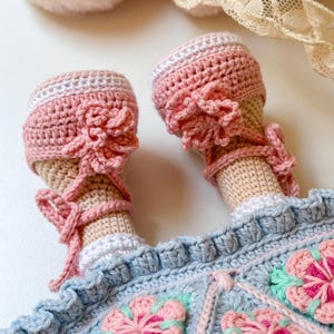 K&ouml;nnte beinhalten: Eine geh&auml;kelte Puppe, die ein blaues Kleid mit rosa Blumen und rosa geh&auml;kelte Schuhe tr&auml;gt.
