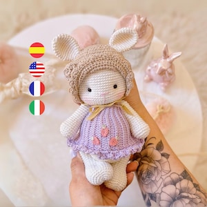 以下が含まれることがあります： 紫色のドレスを着て、刺繍されたイチゴと茶色の編み物帽子をかぶった、編み物で作られたウサギのぬいぐるみ。ぬいぐるみは、腕に花のタトゥーをした人が持っています。