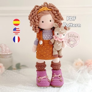 Puede incluir: Una muñeca de ganchillo hecha a mano con cabello rizado castaño, que lleva un mono naranja, un suéter morado y botas rosas. La muñeca sostiene un pequeño ciervo de ganchillo. La imagen incluye el texto "PDF Pattern" y un logotipo.