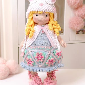 K&ouml;nnte beinhalten: Eine geh&auml;kelte Puppe mit einem wei&szlig;en Hasenhut, einem blauen und rosa Kleid und rosa Schuhen. Die Puppe hat blonde lockige Haare und ein rosa Band um den Hals.