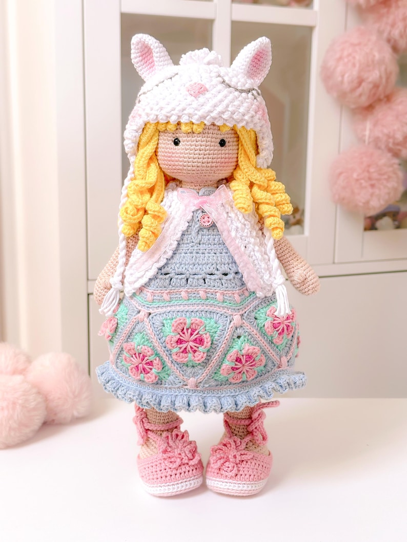 K&ouml;nnte beinhalten: Eine geh&auml;kelte Puppe mit einem wei&szlig;en Hasenhut, einem blauen und rosa Kleid und rosa Schuhen. Die Puppe hat blonde lockige Haare und ein rosa Band um den Hals.