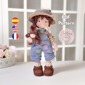 Pode incluir: Uma boneca de crochê com chapéu cinza, camisa listrada e jardineira azul com remendos, segurando um pequeno cavalo de brinquedo marrom. A boneca tem cabelo castanho encaracolado e sapatos marrons. Texto PDF Pattern e Sutitas.