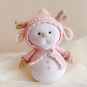 Puede incluir: Un mu&ntilde;eco de nieve de crochet que lleva un su&eacute;ter rosa con capucha y cuernos. El mu&ntilde;eco tiene un bot&oacute;n con un dise&ntilde;o floral en la parte delantera del su&eacute;ter.