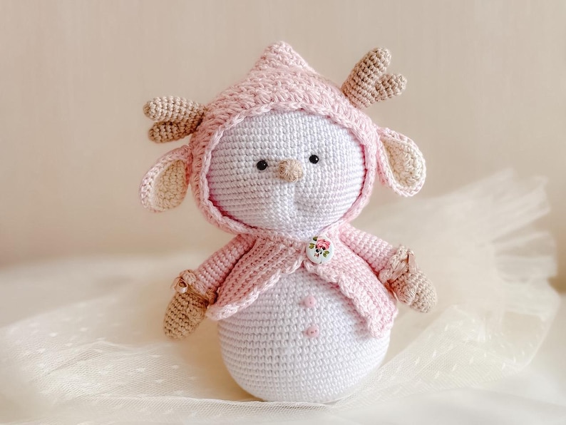 Puede incluir: Un mu&ntilde;eco de nieve de crochet que lleva un su&eacute;ter rosa con capucha y cuernos. El mu&ntilde;eco tiene un bot&oacute;n con un dise&ntilde;o floral en la parte delantera del su&eacute;ter.