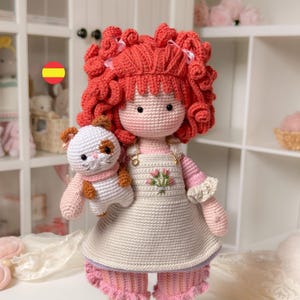 Könnte beinhalten: Handgemachte Häkelpuppe mit roten Locken und einem rosa-weißen Outfit, die eine kleine Häkelkatze hält. Das Kleid der Puppe hat ein Blumenmuster. Die Puppe ist etwa 30 cm groß.