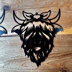 May include: Black metal cutout of a Highland cow head with a bow. The cow has a fluffy mane and a detailed face.