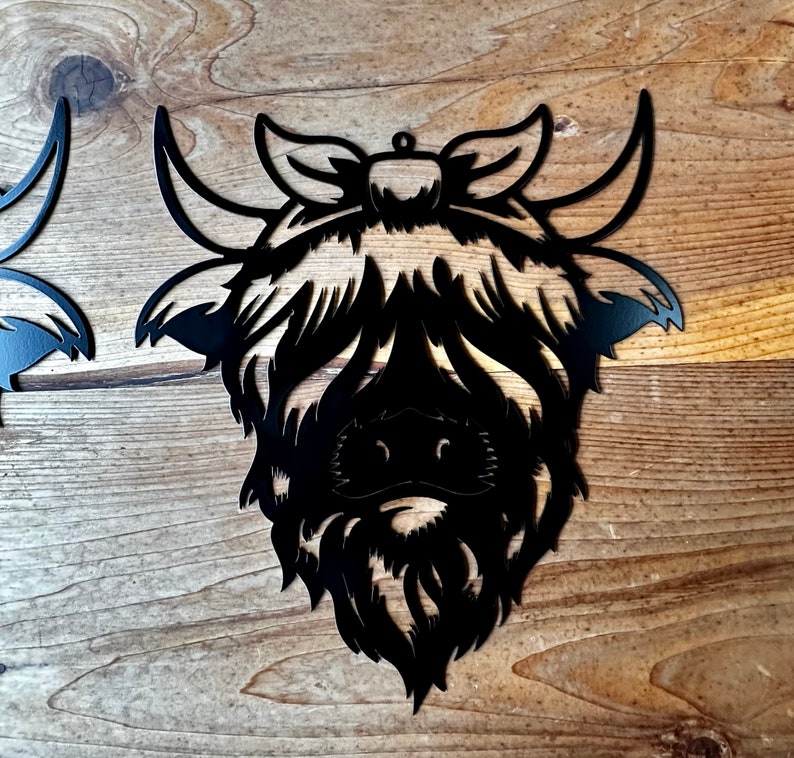 May include: Black metal cutout of a Highland cow head with a bow. The cow has a fluffy mane and a detailed face.