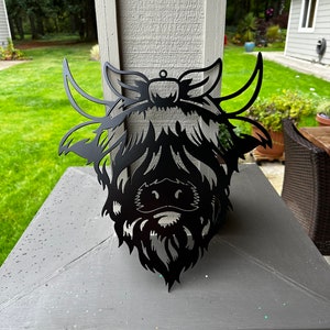 May include: Black metal silhouette of a Highland cow head with a long beard and large horns. The silhouette is mounted on a gray stone surface.