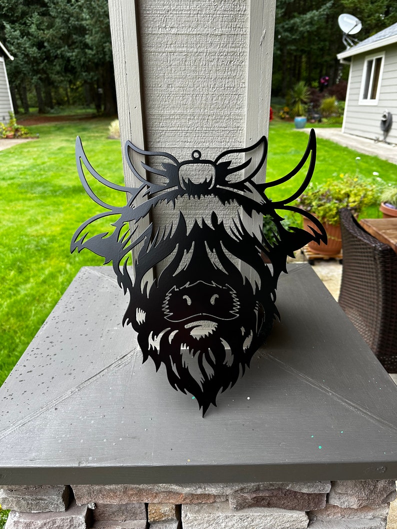 May include: Black metal silhouette of a Highland cow head with a long beard and large horns. The silhouette is mounted on a gray stone surface.