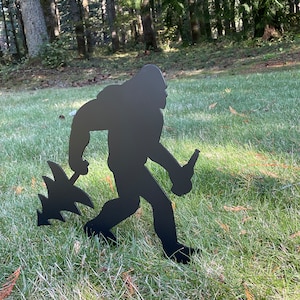 May include: Black metal silhouette of a Bigfoot holding a beer bottle and a small tree in a grassy area.