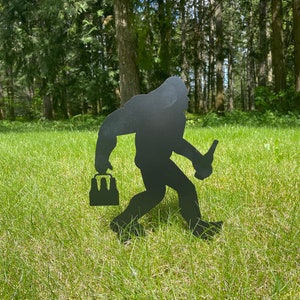 May include: Black metal silhouette of a Bigfoot carrying a six-pack of beer and holding a beer bottle in his other hand. The silhouette is standing on a green grassy lawn.