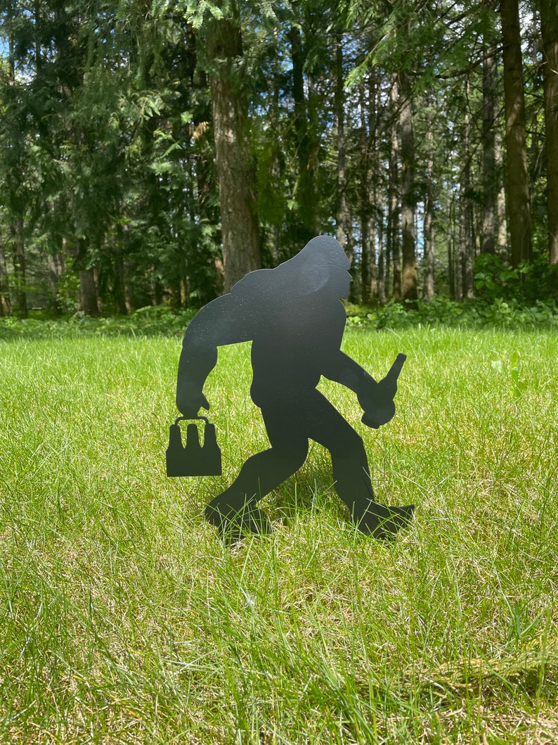 May include: Black metal silhouette of a Bigfoot carrying a six-pack of beer and holding a beer bottle in his other hand. The silhouette is standing on a green grassy lawn.