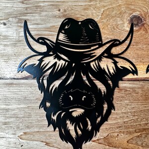 May include: Black metal silhouette of a Highland cow wearing a cowboy hat. The cow's face is detailed with a beard and horns.