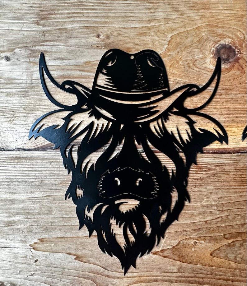 May include: Black metal silhouette of a Highland cow wearing a cowboy hat. The cow's face is detailed with a beard and horns.