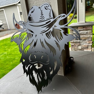 May include: Black metal silhouette of a Highland cow wearing a cowboy hat. The cow's head is facing forward with its horns pointing up and out. The hat has a wide brim and a band around the crown.