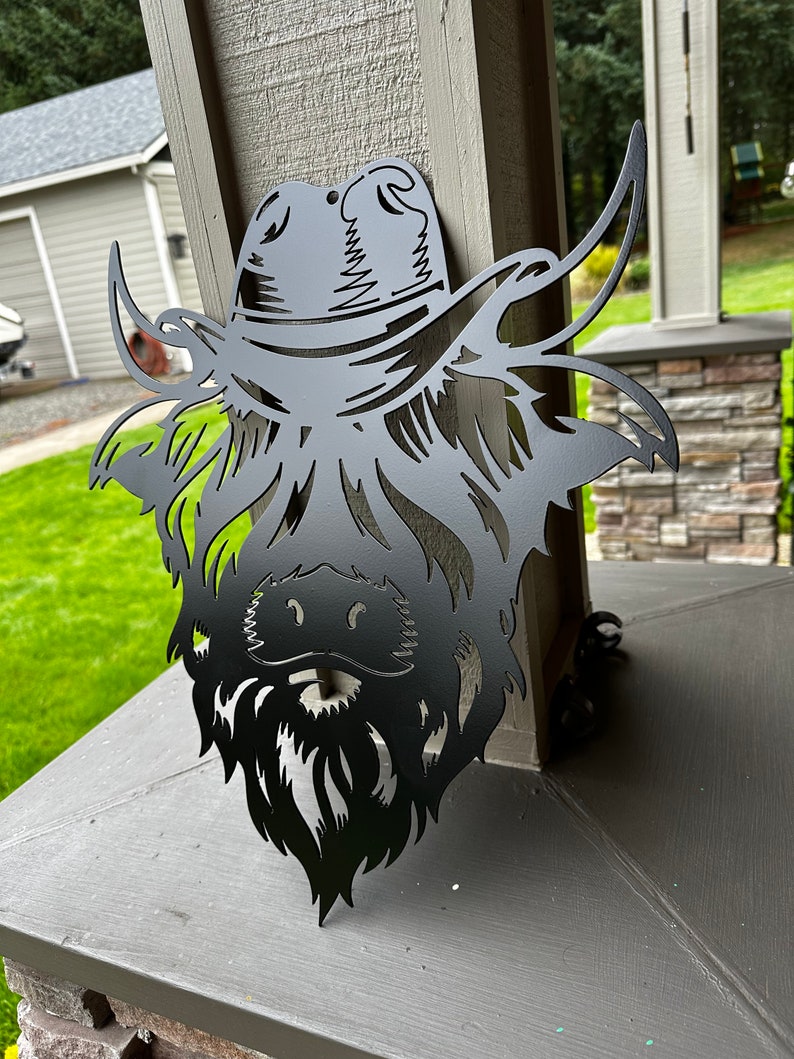 May include: Black metal silhouette of a Highland cow wearing a cowboy hat. The cow's head is facing forward with its horns pointing up and out. The hat has a wide brim and a band around the crown.