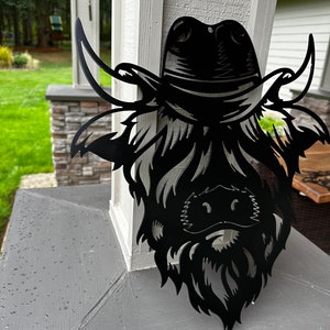 May include: Black metal wall art depicting a Highland cow wearing a cowboy hat. The cow's face is detailed with a beard and horns.