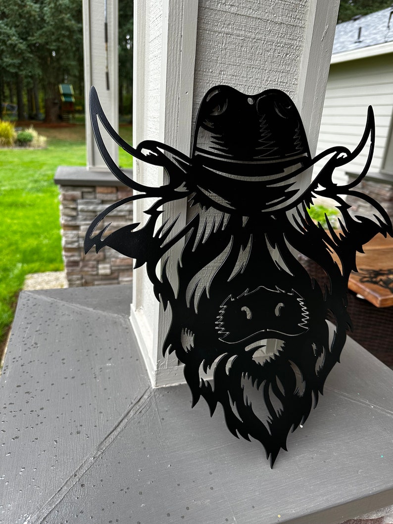 May include: Black metal wall art depicting a Highland cow wearing a cowboy hat. The cow's face is detailed with a beard and horns.