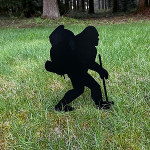 May include: Black metal silhouette of a person hiking with a backpack and a walking stick. The silhouette is standing on a green grassy lawn.