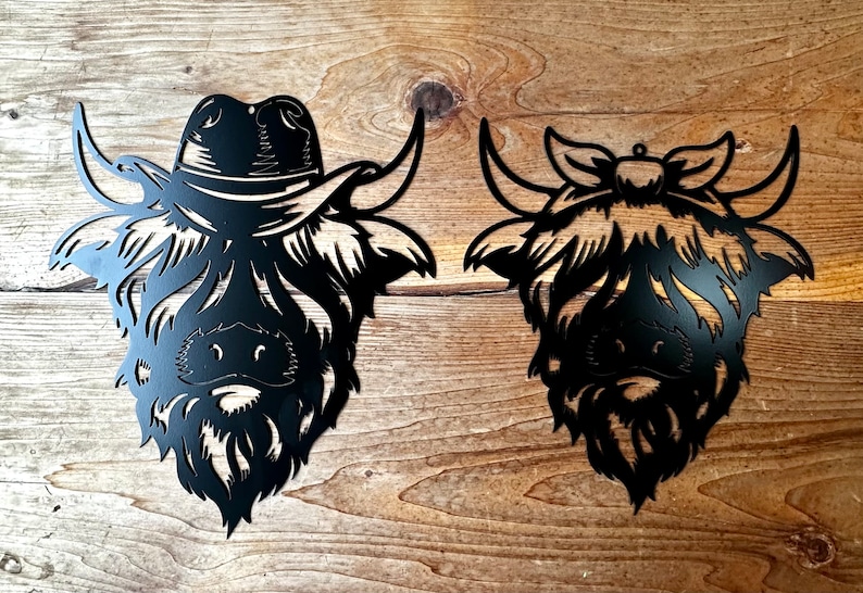 May include: Two black metal Highland Cow head silhouettes, one wearing a cowboy hat and the other a bow. The silhouettes are mounted on a wooden surface.