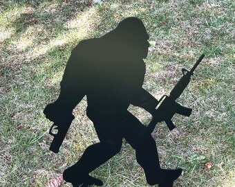 May include: Black metal silhouette of a Bigfoot holding a rifle in a walking pose. The silhouette is standing on a green grassy lawn.