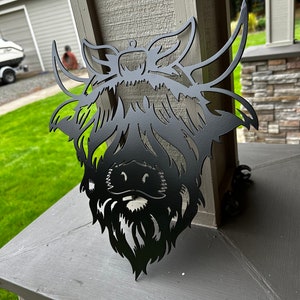 May include: Black metal silhouette of a Highland cow head with a long, shaggy mane and beard. The silhouette is mounted on a gray surface.