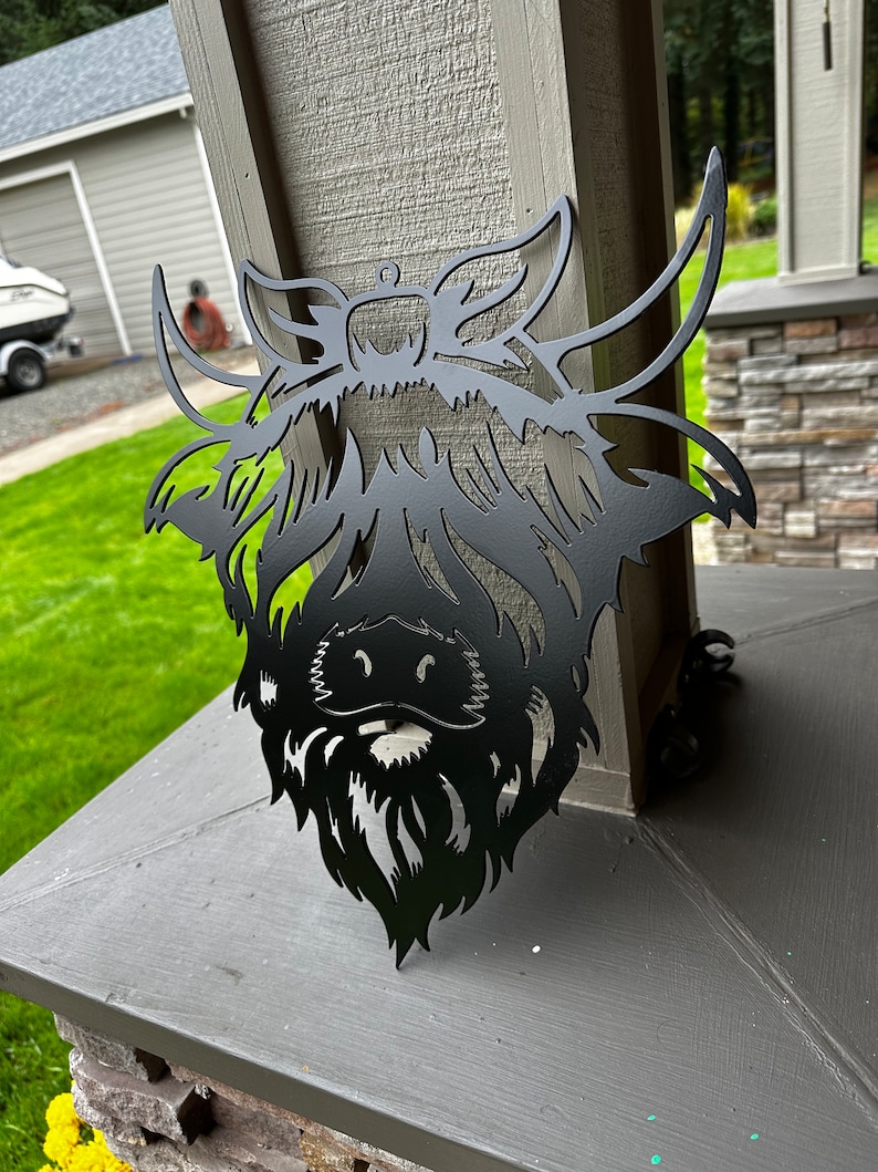 May include: Black metal silhouette of a Highland cow head with a long, shaggy mane and beard. The silhouette is mounted on a gray surface.