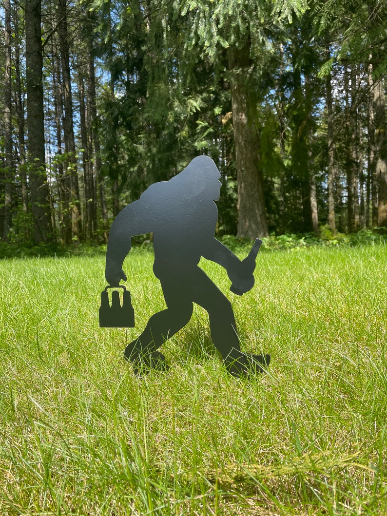 May include: A black metal silhouette of a Bigfoot carrying a six-pack of beer and a bottle in one hand. The silhouette is standing in a grassy field with trees in the background.
